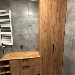 Dawid Brociek - Drewniana szafka łazienkowa z szufladami i otwartymi półkami, obok wysoka szafa z dwoma drzwiami na tle szarych płytek. Nowoczesny design łazienki.