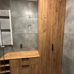 Drewniana szafka łazienkowa z szufladami i otwartymi półkami, obok wysoka szafa z dwoma drzwiami na tle szarych płytek. Nowoczesny design łazienki.