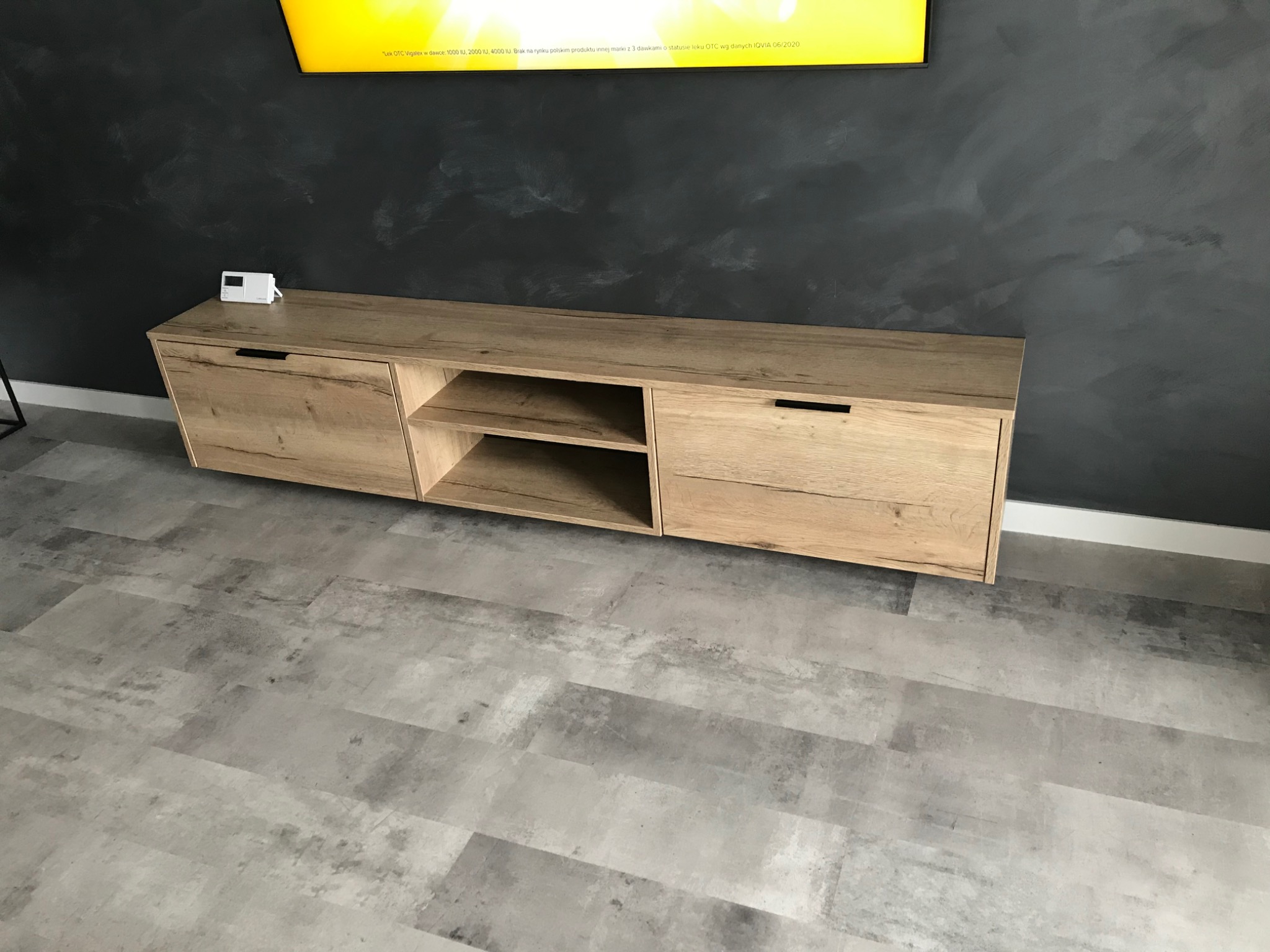 Drewniana szafka RTV w odcieniach dębu, zawieszona na szarej ścianie z efektem betonu, z widocznym fragmentem telewizora u góry i szarą podłogą imitującą kamień.