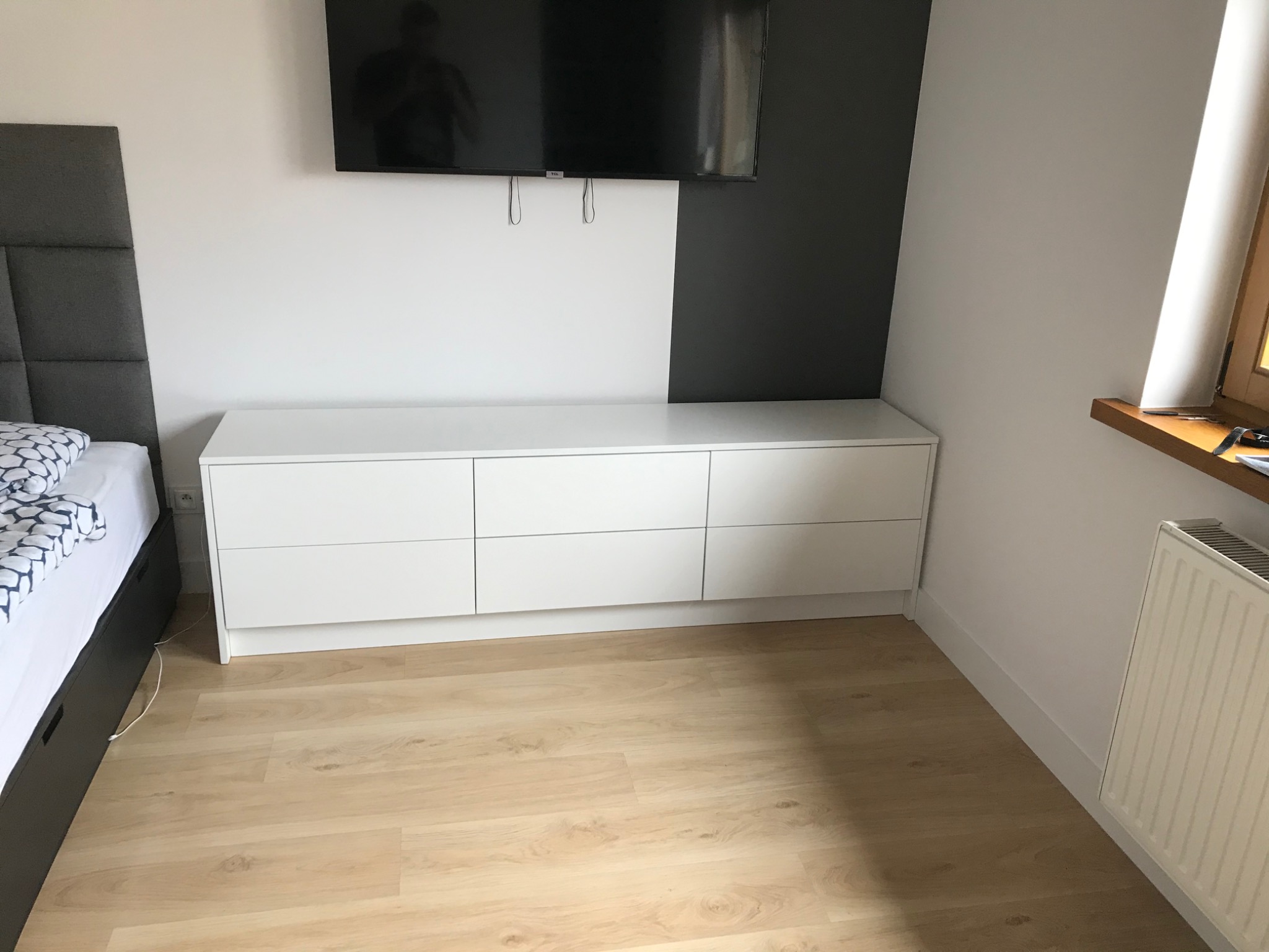 Biała komoda z sześcioma szufladami w sypialni, ustawiona pod telewizorem na ścianie pomalowanej w dwóch kolorach, obok łóżko z szarym zagłówkiem.