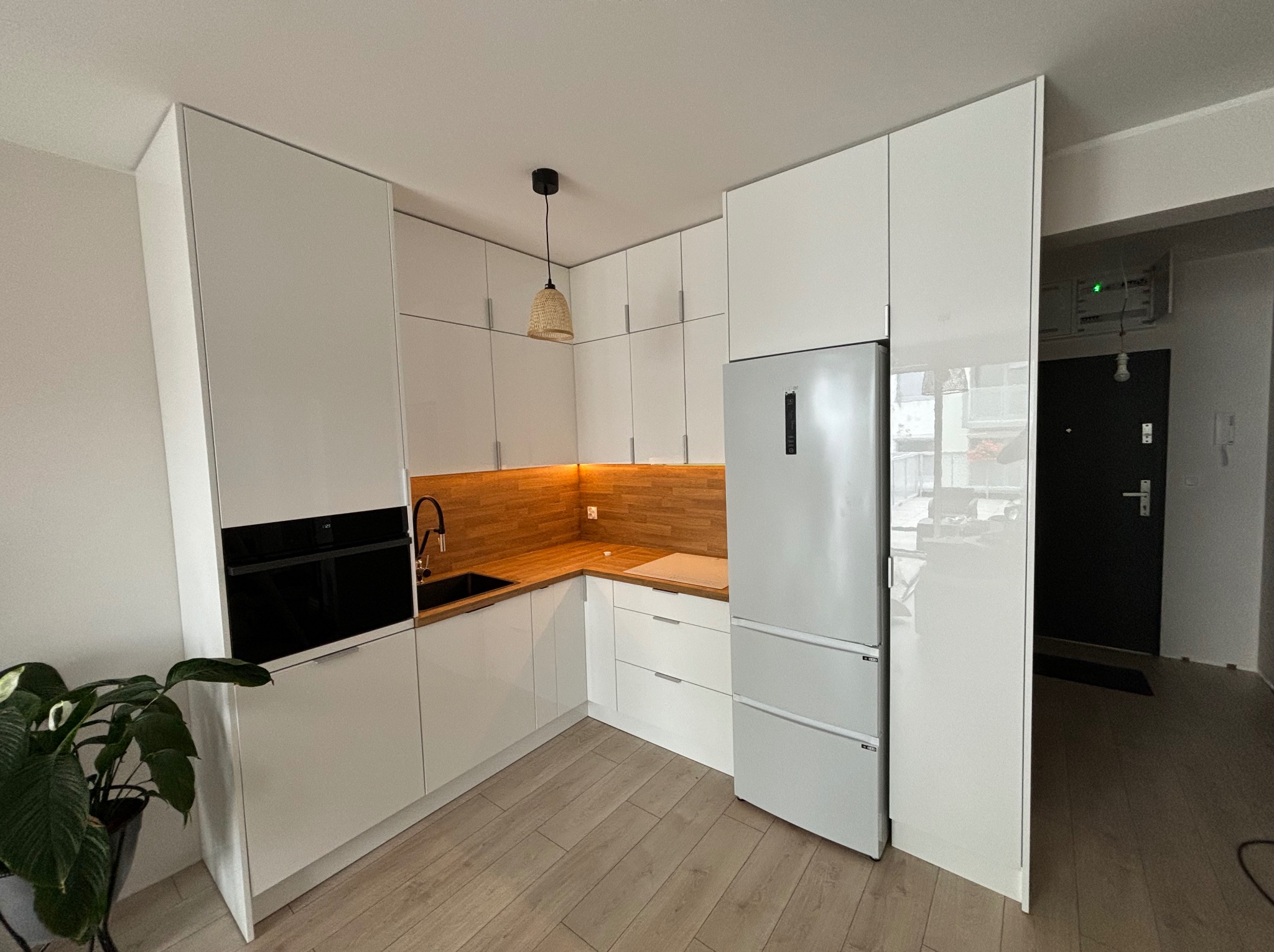 Jasna kuchnia z drewnianym blatem i oświetleniem LED, minimalistyczny design, fronty szafek na wysoki połysk, widok na fragment korytarza z drzwiami.