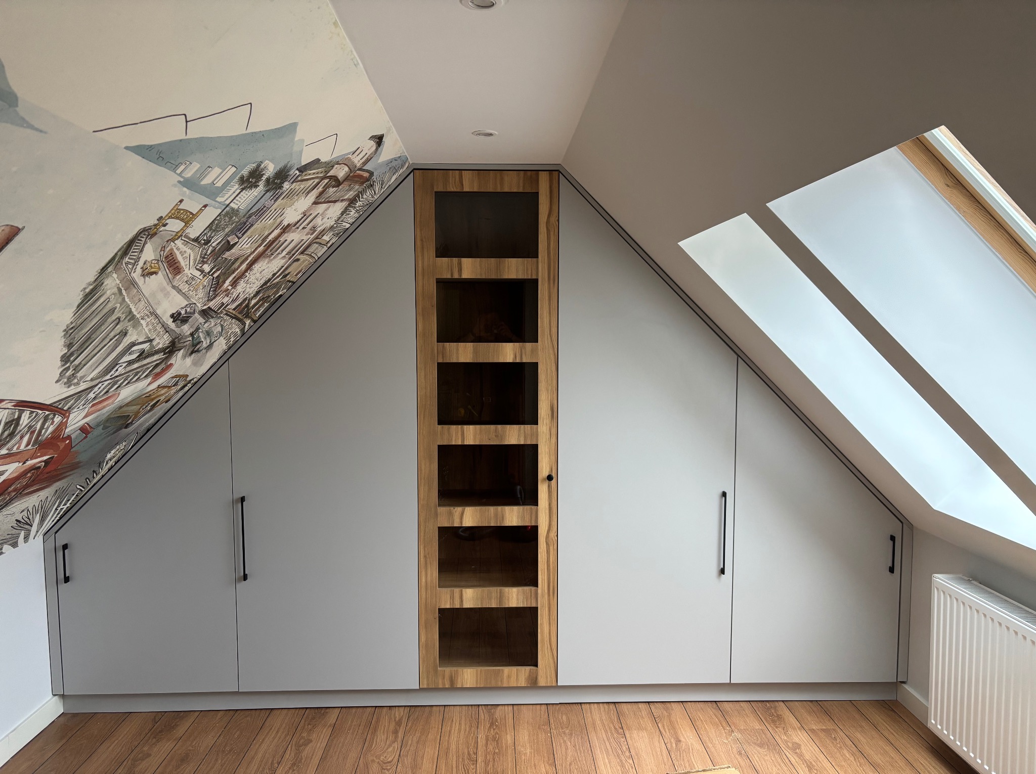 Szafa wnękowa na poddaszu ze ścianą ozdobioną malowidłem. Fronty w kolorze szarym, środkowa część z drewnianą ramą i przeszkleniami. Widoczne okno dachowe.