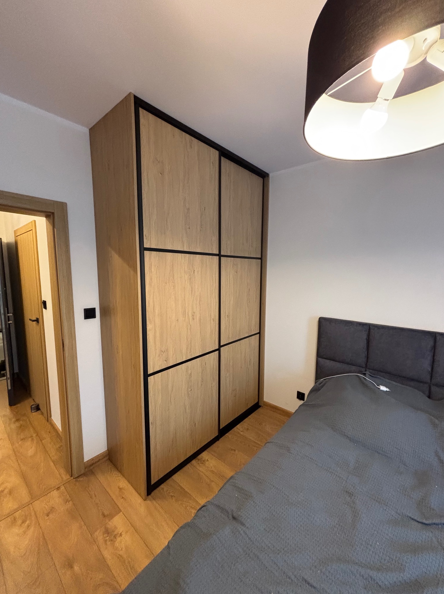 Szafa przesuwna z drewnopodobnymi frontami podzielonymi czarnymi listwami, w sypialni z łóżkiem i drewnianą podłogą. Widok na otwarte drzwi do innego pomieszczenia.