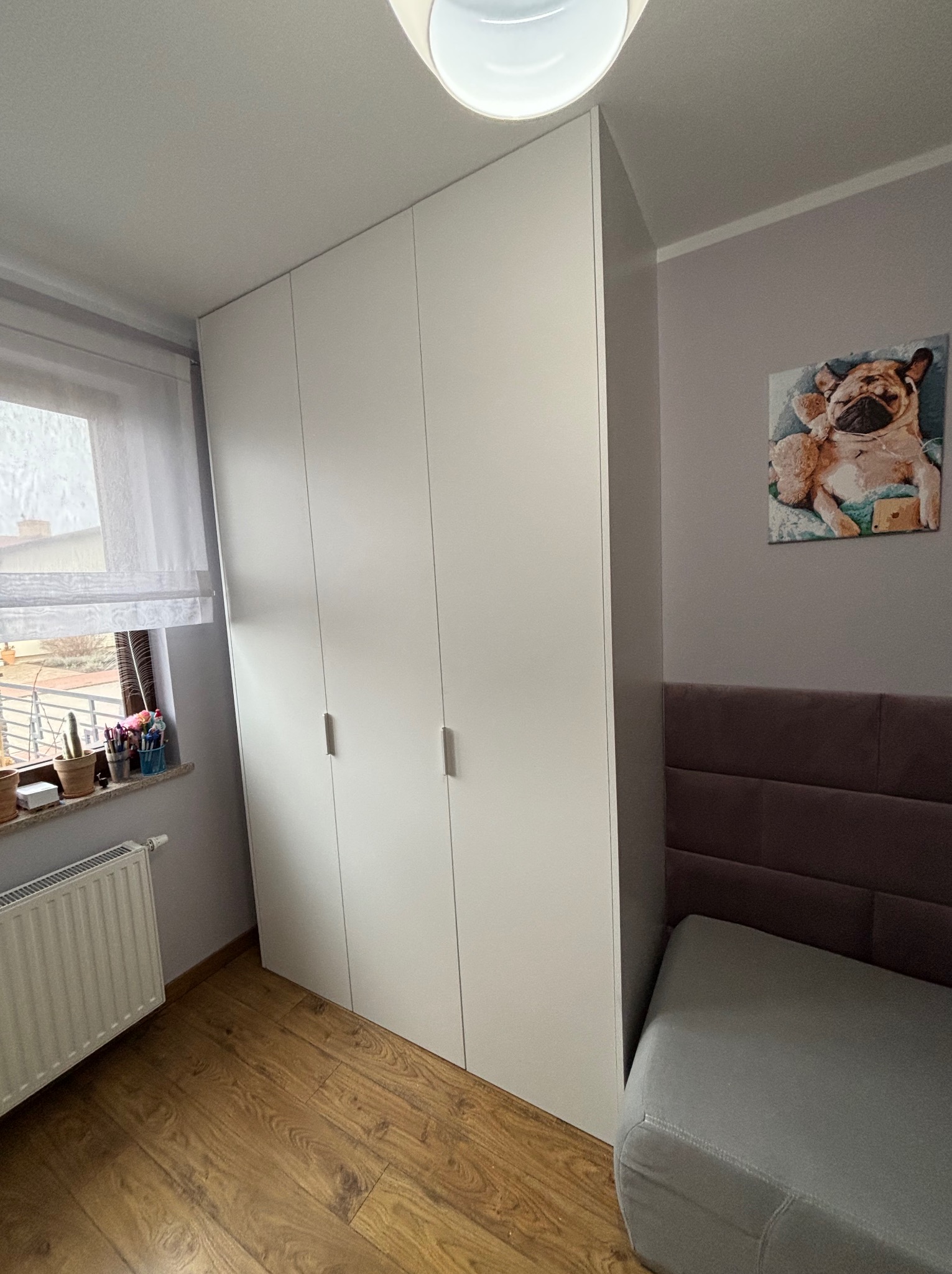 Biała, czterodrzwiowa szafa wnękowa z podłużnymi uchwytami, ustawiona w pokoju z drewnianą podłogą, obok okna i łóżka z wezgłowiem. Na ścianie wisi obrazek.