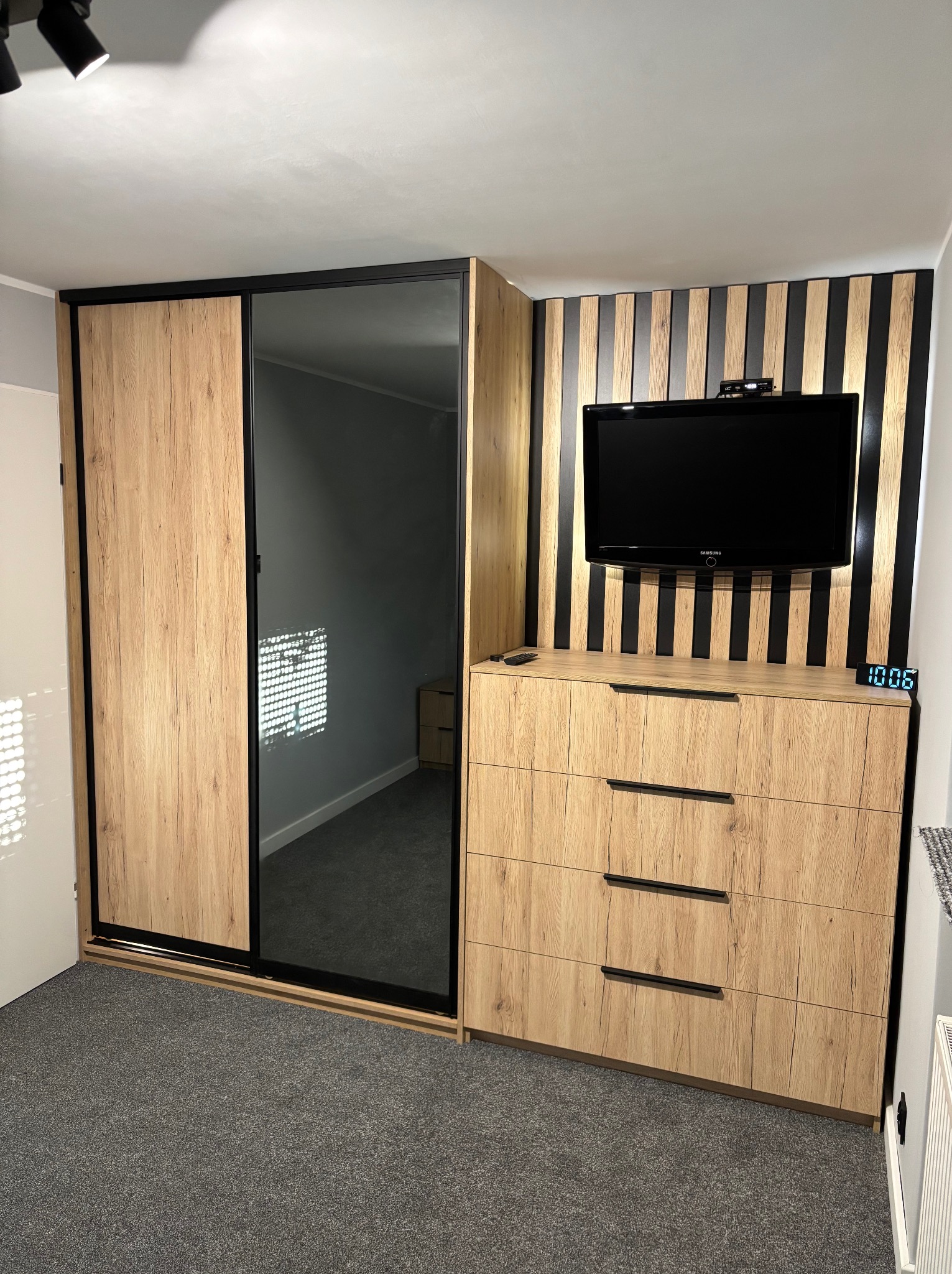 Szafa przesuwna z lustrem i komoda z TV wkomponowana w lamelową ściankę, imitacja drewna, czarne detale. Realizacja w Poznaniu.