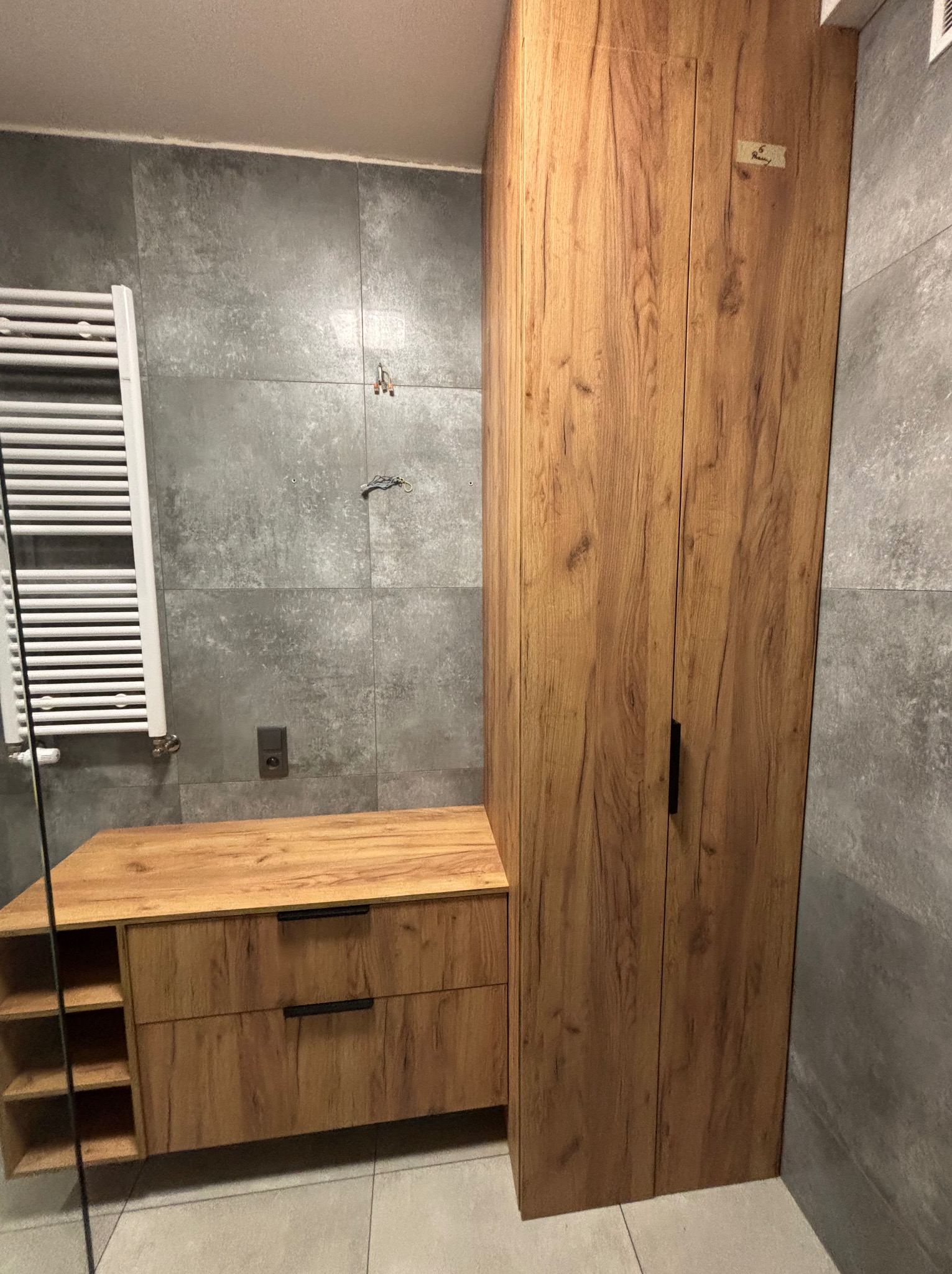 Drewniana szafka łazienkowa z szufladami i otwartymi półkami, obok wysoka szafa z dwoma drzwiami na tle szarych płytek. Nowoczesny design łazienki.