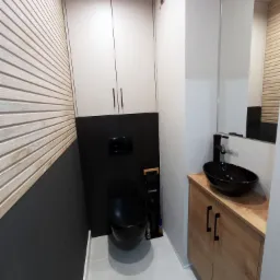 Nowoczesna toaleta z drewnianą boazerią, szafką pod umywalkę z drewnianym blatem i czarną umywalką nablatową, zabudową nad toaletą i czarną ścianą za toaletą.
