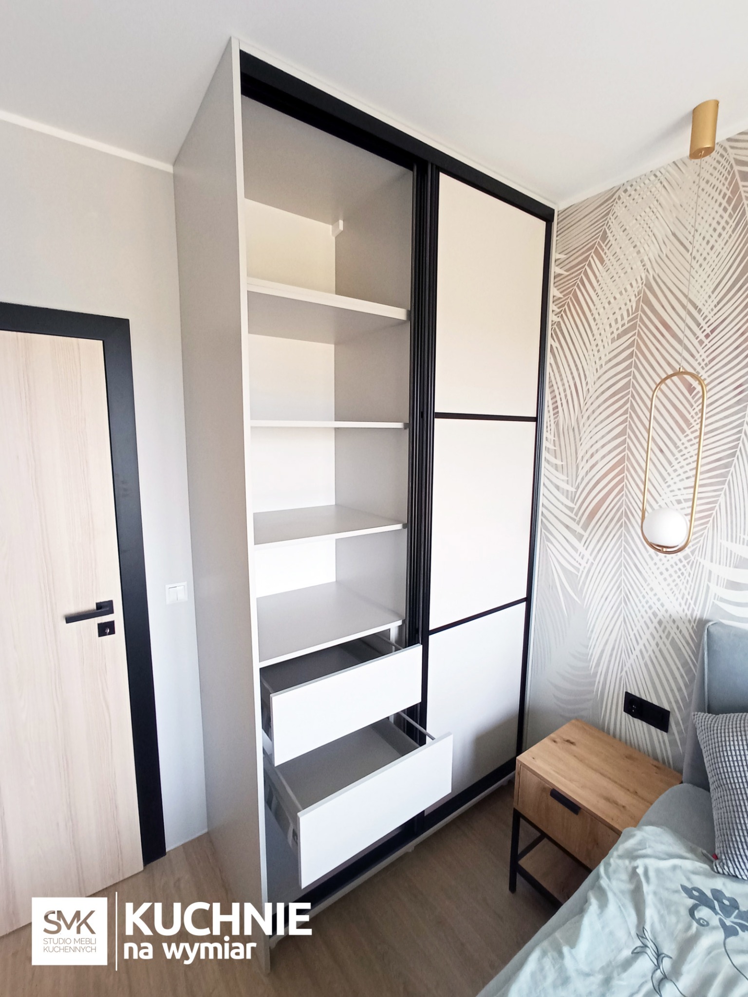 Biała szafa wnękowa z przesuwnymi drzwiami z czarnymi obramowaniami, częściowo otwarta, ukazująca półki i wysuwane szuflady, w sypialni z tapetą w liście i drewnianą szafką nocną.