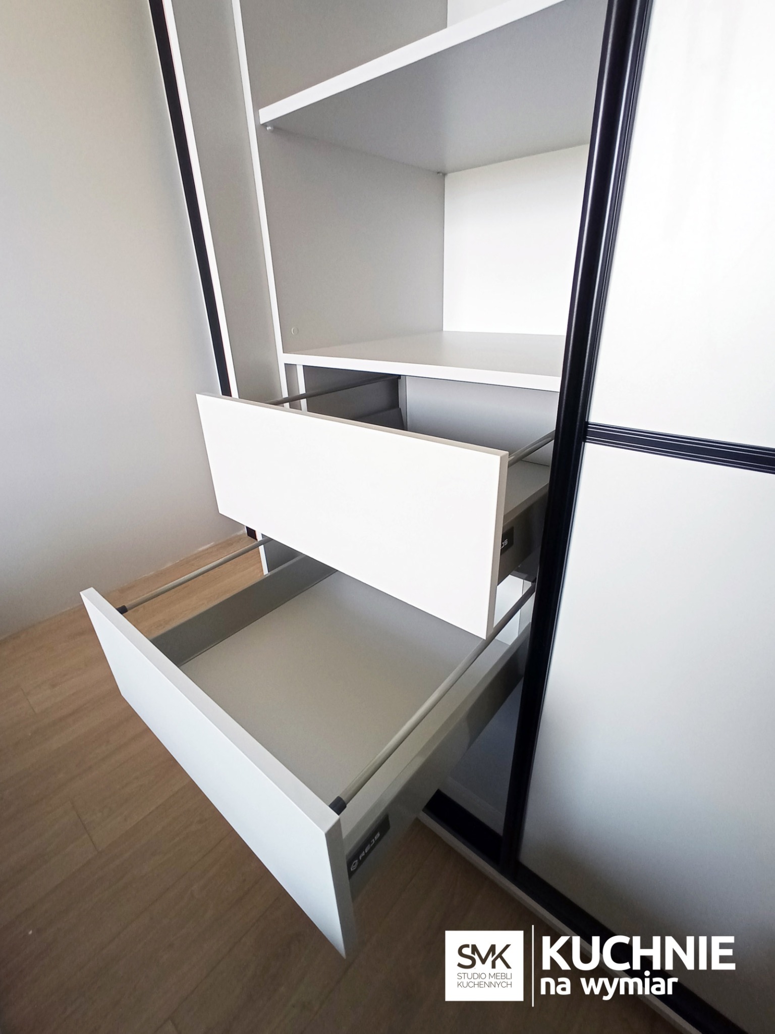 Wnętrze szafy wnękowej z otwartymi, białymi szufladami na prowadnicach, widoczne półki i czarne ramy drzwi przesuwnych.