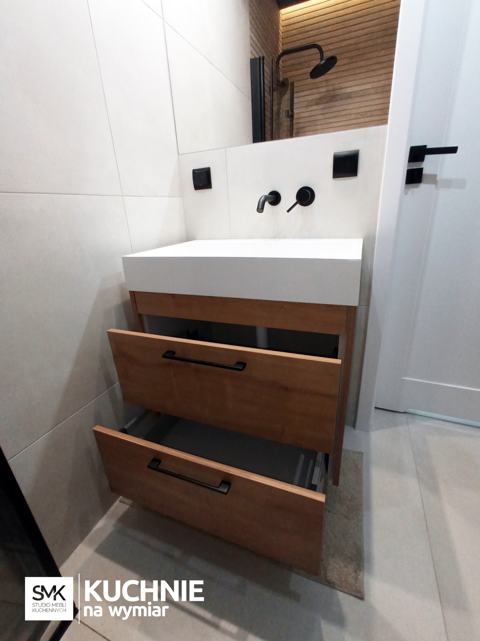 Nowoczesna szafka łazienkowa z dwoma wysuniętymi szufladami z jasnego drewna z czarnymi, długimi uchwytami, biały blat umywalki, czarna bateria ścienna i włączniki na tle jasnych płytek i fragmentu...