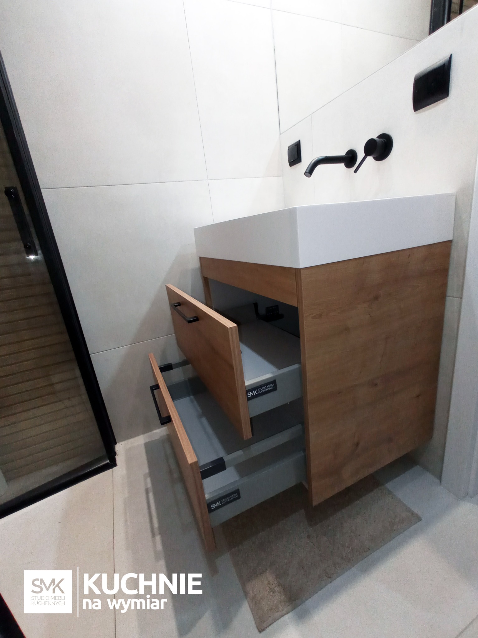 Nowoczesna szafka łazienkowa pod umywalkę z dwoma wysuniętymi szufladami, drewnopodobny fornir, biały blat, czarna armatura ścienna, minimalistyczny design.