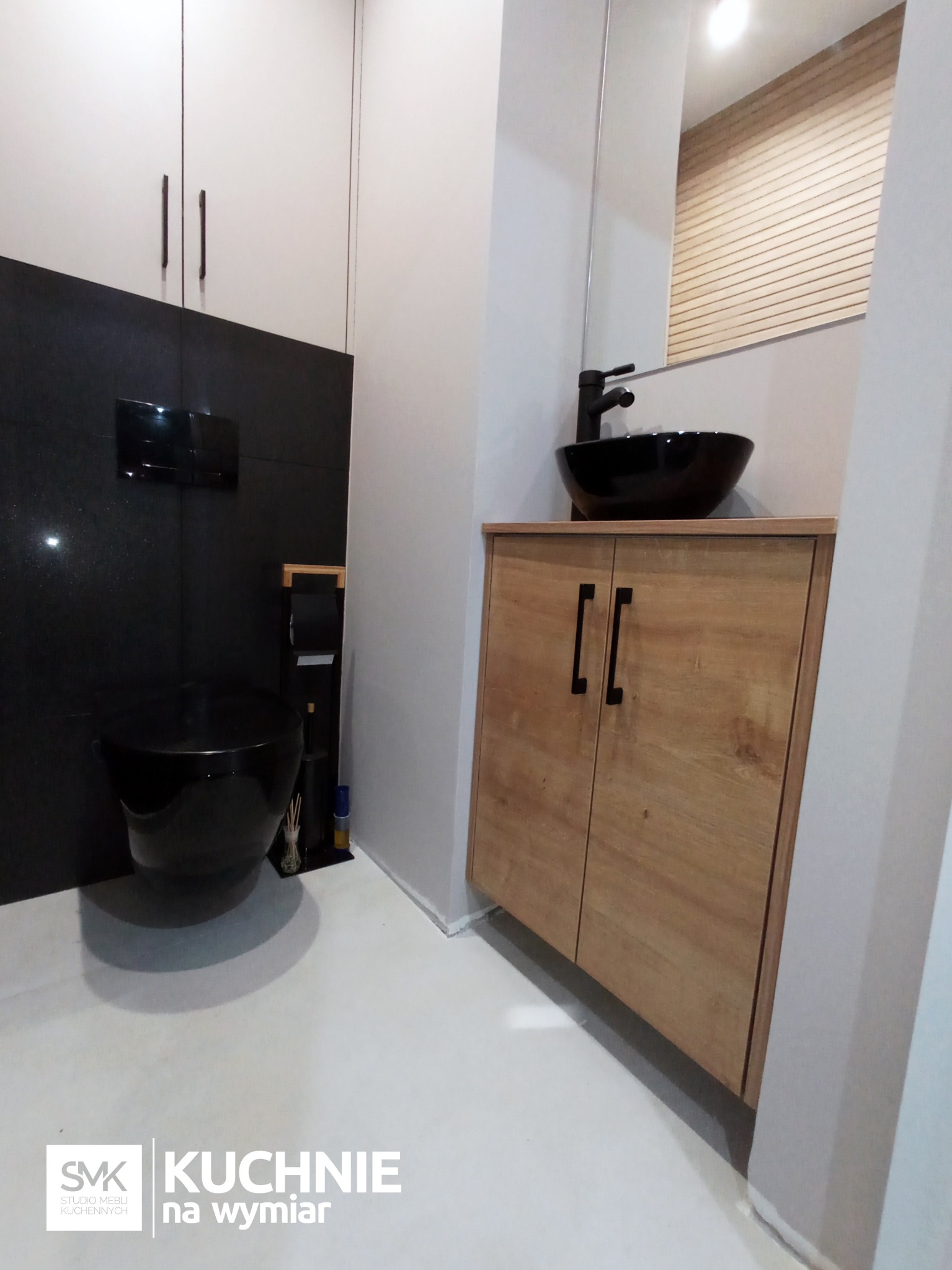 Nowoczesna łazienka z czarną ceramiką: wisząca toaleta i umywalka na drewnianej szafce z czarnymi uchwytami, na tle białych i czarnych płytek oraz jasnej podłogi.