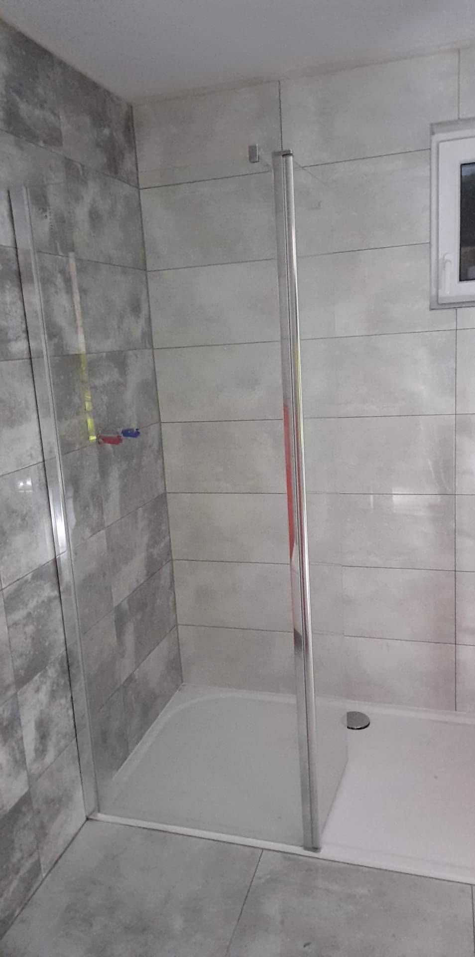 Szara łazienka z kabiną prysznicową typu walk-in, wykończona płytkami imitującymi beton, z widocznymi przyłączami wodnymi.