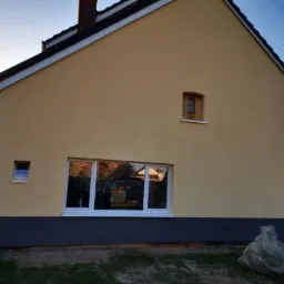 Żółta elewacja domu jednorodzinnego z widoczną szarą podmurówką, białymi oknami i małym drewnianym okienkiem na poddaszu, uchwycona w popołudniowym świetle.