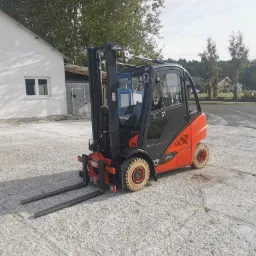 Pomarańczowo-czarny wózek widłowy Linde H20 z kabiną, stojący na betonowej powierzchni, z białym budynkiem i drzewami w tle.