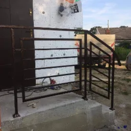 Metalowa konstrukcja schodów zewnętrznych z poręczą w trakcie montażu, widoczne narzędzia i elementy budynku w tle.