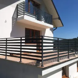 Nowoczesna, pozioma, czarna balustrada metalowa na balkonie domu z białymi ścianami i brązowymi oknami, widok z dołu w słoneczny dzień.