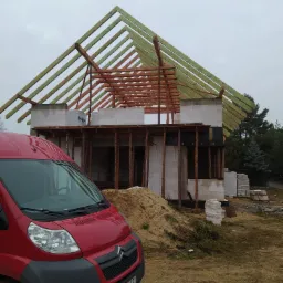 Budowa domu w trakcie realizacji: surowy stan otwarty z widoczną konstrukcją drewnianą dachu, ściany z bloczków betonowych H+H, czerwony samochód dostawczy Citroen na pierwszym planie, plac budowy.