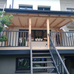 ASW-DACH Adrian Wierowicz - Nowo wybudowany taras z drewnianą konstrukcją zadaszenia, czarnymi balustradami i schodami, widok z dołu, drabina oparta o budynek obok drzewa.