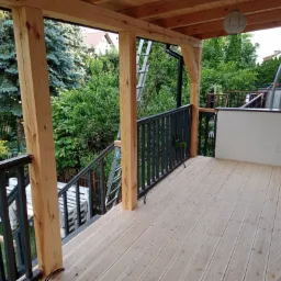 Nowy taras drewniany z balustradą, widok z góry na deski podłogowe, słupy konstrukcyjne i metalowe barierki, drabina oparta o konstrukcję w tle.