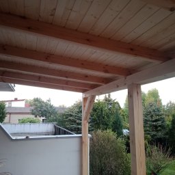 ASW-DACH Adrian Wierowicz - Drewniana konstrukcja z belek i desek, prawdopodobnie zadaszenie tarasu, w trakcie budowy, widok od dołu, na zewnątrz zieleń drzew i krzewów.