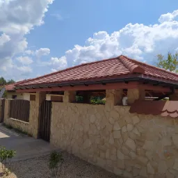 Nowoczesny dom z czerwoną dachówką karpiówką, kamiennym ogrodzeniem i widoczną drabiną opartą o ścianę, na tle błękitnego nieba z chmurami.