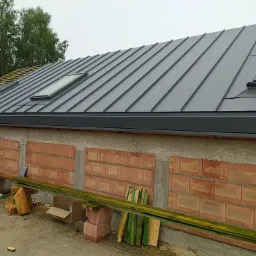 Szary dach z blachy na budynku w trakcie budowy, widoczne okna dachowe i fragment więźby dachowej bez pokrycia, na dole mur z cegły dziurawki.