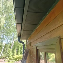 Szary system rynnowy zamontowany na drewnianej konstrukcji budynku z widoczną drabinką opartą o ścianę i zielonym ogrodem w tle.