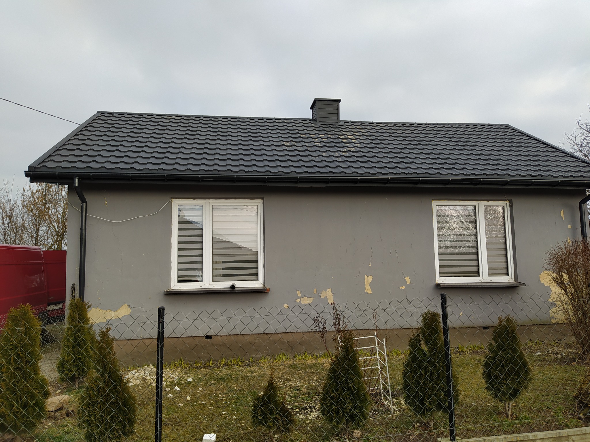 Dom jednorodzinny z nowym, ciemnoszarym dachem z blachodachówki, widoczny komin i rynny, elewacja w trakcie renowacji z widocznymi odpadającymi fragmentami farby, ogrodzenie z siatki.