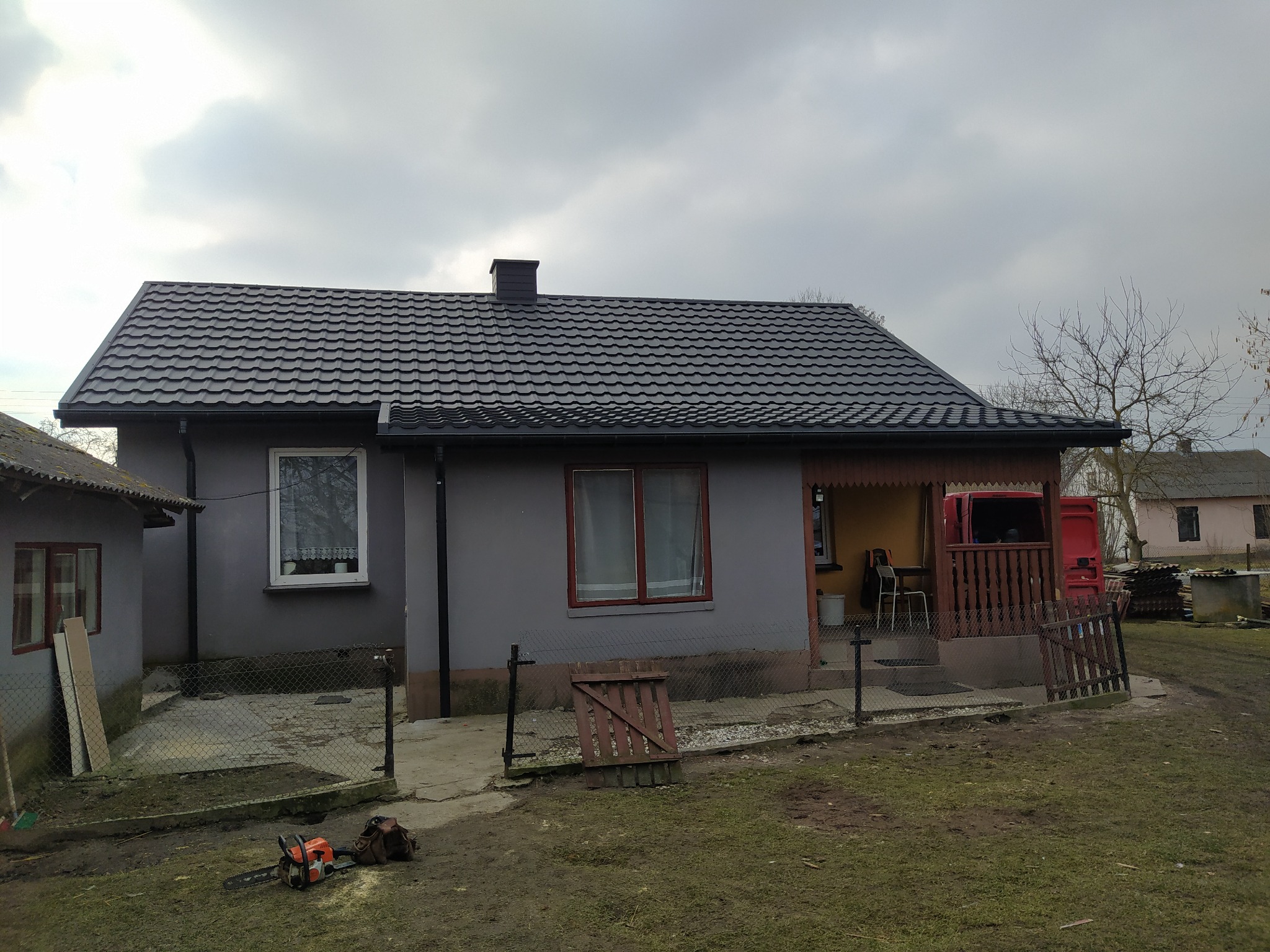 Dom jednorodzinny z nowym, ciemnoszarym dachem z blachodachówki, widoczne obróbki blacharskie, komin i rynny. Na pierwszym planie piła spalinowa i teren po wycince drzew.