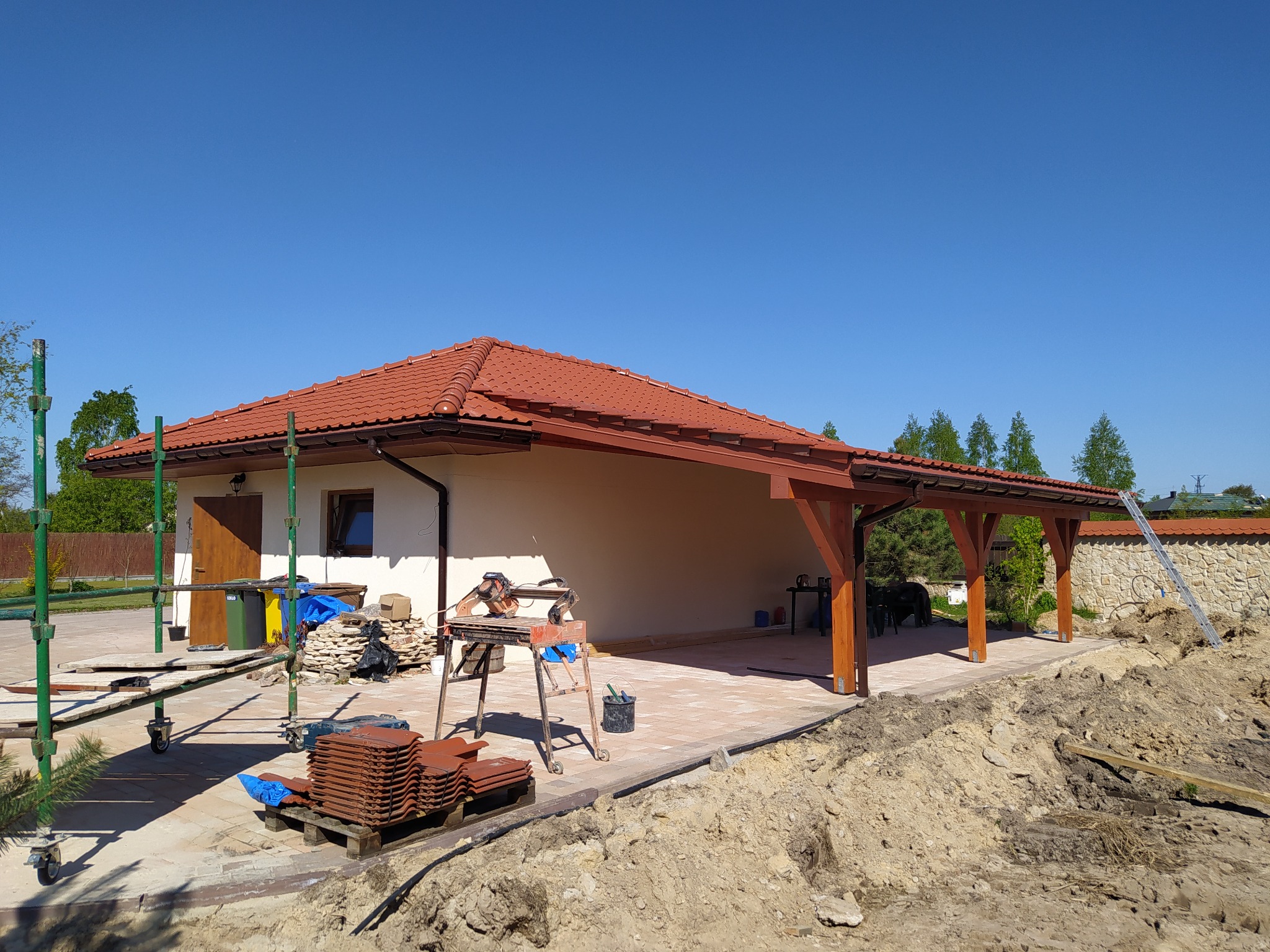 Budynek z nowym dachem z czerwonej dachówki ceramicznej i drewnianą konstrukcją zadaszenia tarasu, widoczna sterta dachówek i piła do cięcia płytek na placu budowy, w tle błękitne niebo.