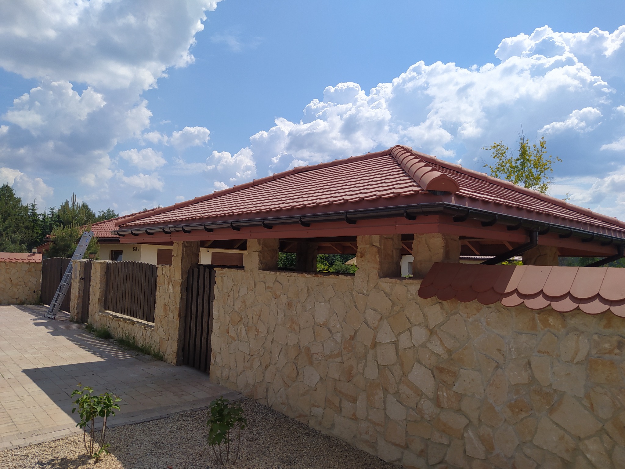 Nowoczesny dom z czerwoną dachówką karpiówką, kamiennym ogrodzeniem i widoczną drabiną opartą o ścianę, na tle błękitnego nieba z chmurami.