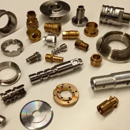 Zestaw różnorodnych, precyzyjnie wykonanych metalowych elementów i komponentów, w tym gwintowane złączki, tuleje, pierścienie i wałki, ułożonych na jasnym tle, prezentujących różne etapy obróbki...