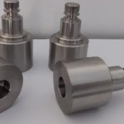 Cztery metalowe elementy obrobione na tokarce lub CNC, dwa stojące pionowo, dwa leżące poziomo, widoczne gwinty i otwory, na białym tle.