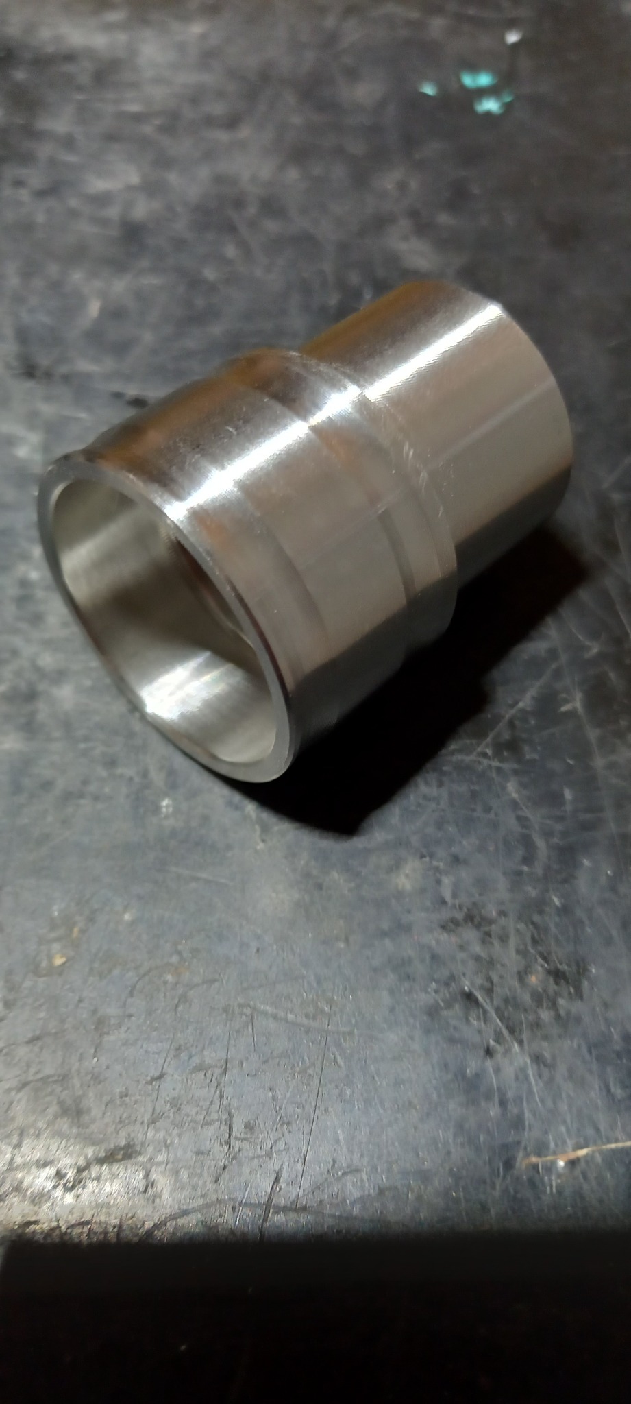 Metalowy, obrobiony element cylindryczny z widocznymi śladami obróbki, leżący na ciemnym, zarysowanym blacie.