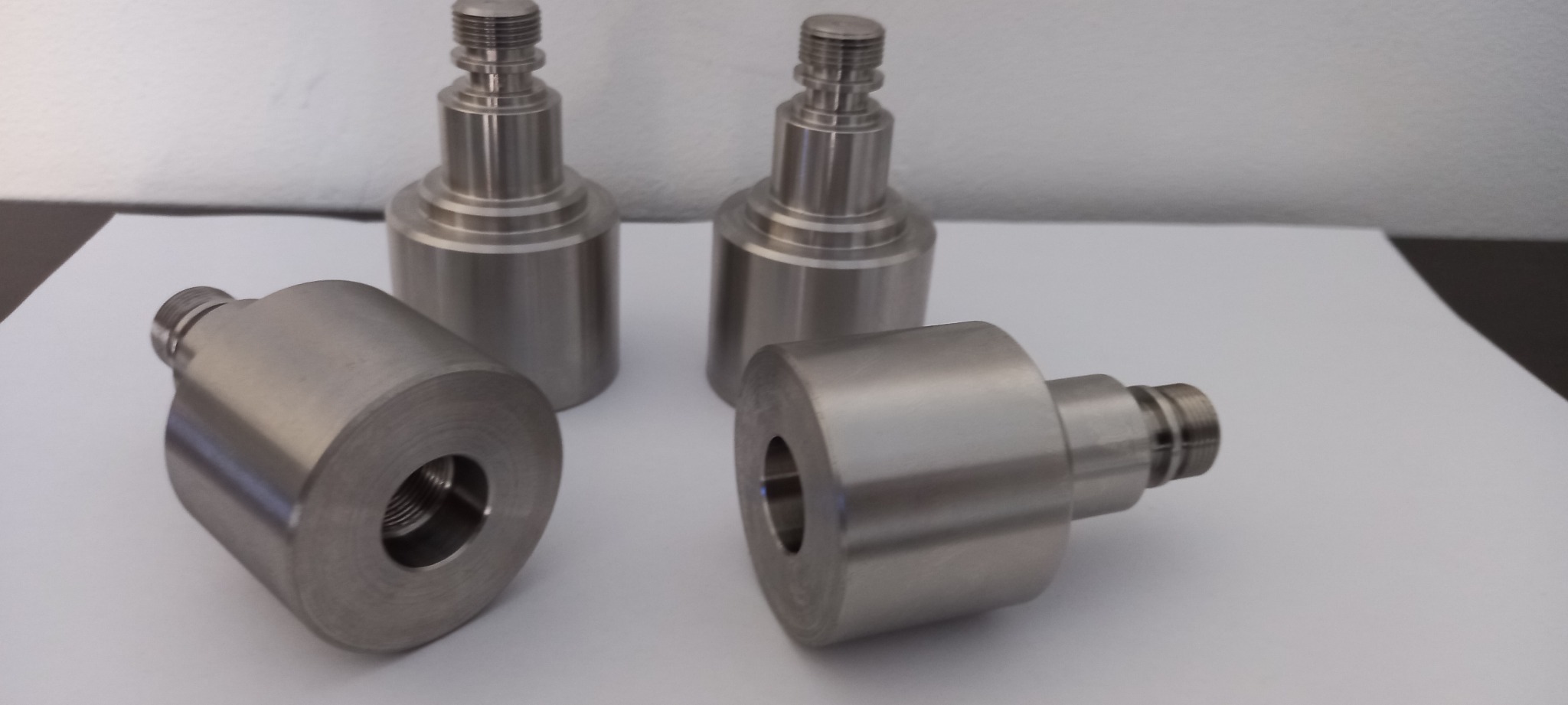 Cztery metalowe elementy obrobione na tokarce lub CNC, dwa stojące pionowo, dwa leżące poziomo, widoczne gwinty i otwory, na białym tle.