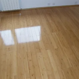 Jasnobrązowa podłoga z paneli, ułożona wzdłuż pokoju, z widocznym odbiciem światła słonecznego w centralnej części, listwy przypodłogowe w kolorze podłogi, białe ściany i grzejnik w tle.