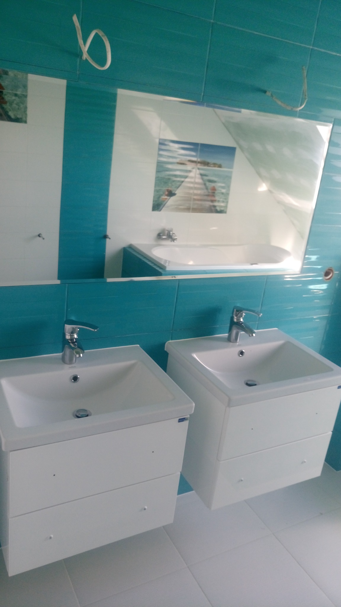 Dwie białe umywalki z szafkami w łazience z turkusowymi płytkami, odbicie w lustrze ukazuje wannę z hydromasażem i dekoracyjną płytkę z motywem plaży.