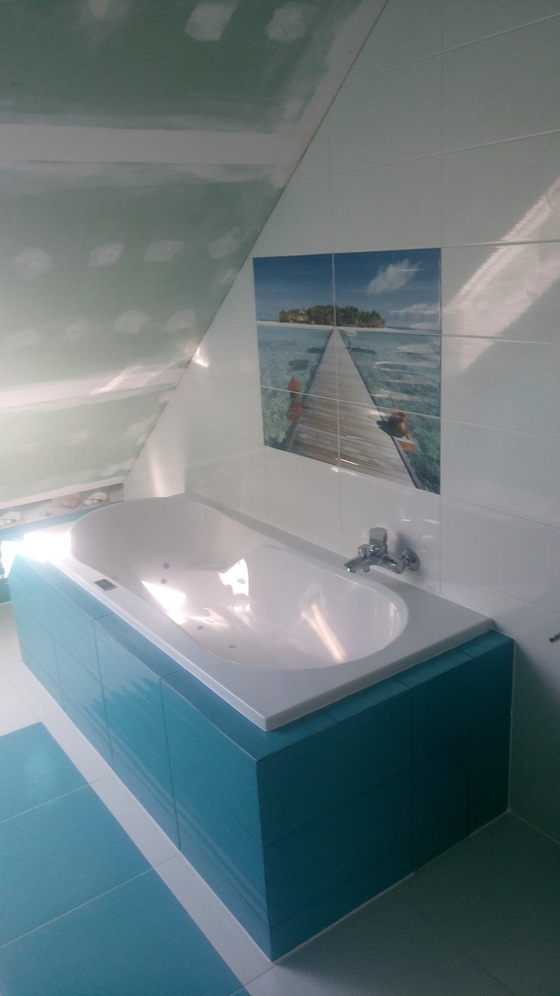 Wanna obudowana turkusowymi płytkami w łazience na poddaszu, ściany wyłożone białymi płytkami z dekoracyjnym motywem morskiego pomostu, sufit częściowo wykończony płytami gipsowo-kartonowymi przed...