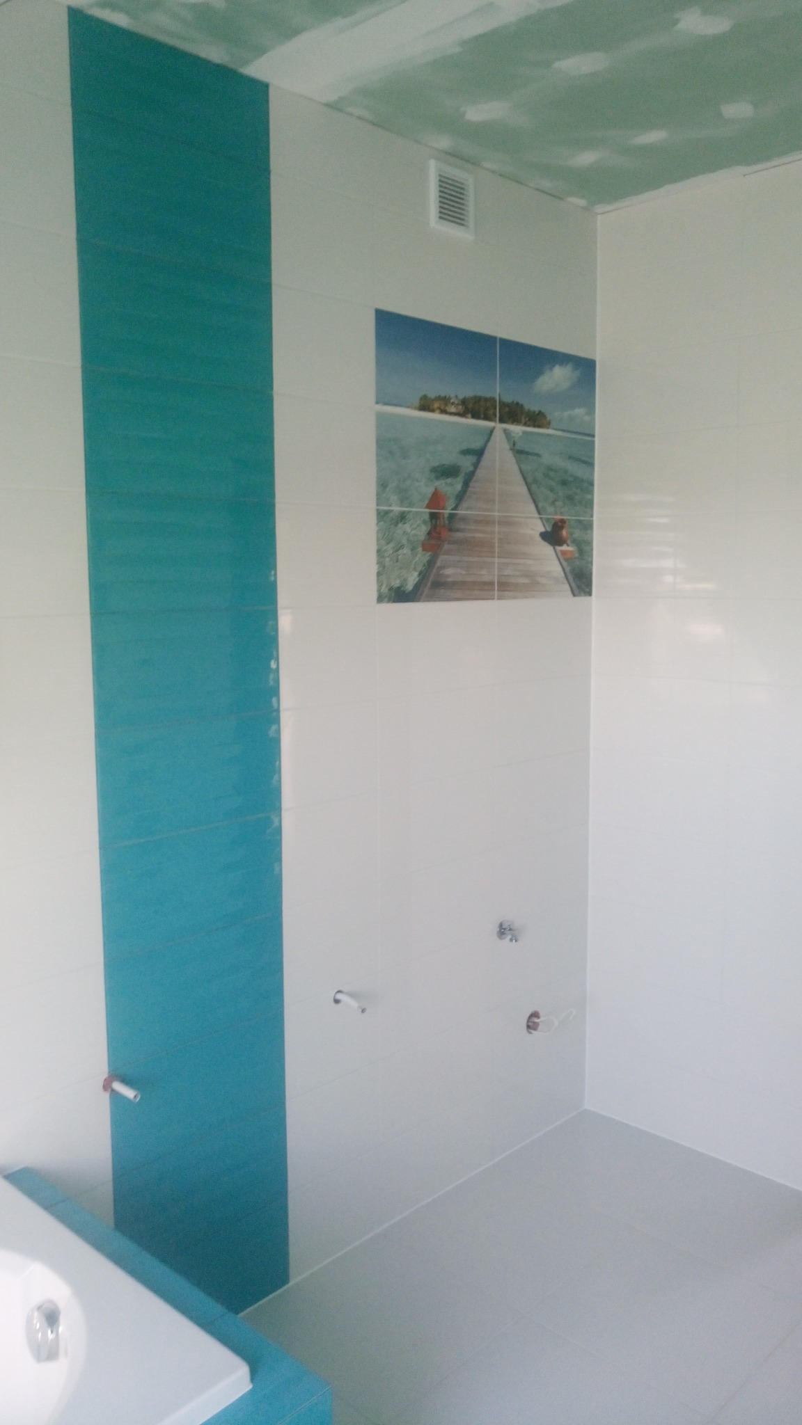 Biało-turkusowa łazienka z fototapetą przedstawiającą molo na tropikalnej wyspie, widoczne niedokończone prace na suficie i przygotowane punkty instalacyjne.