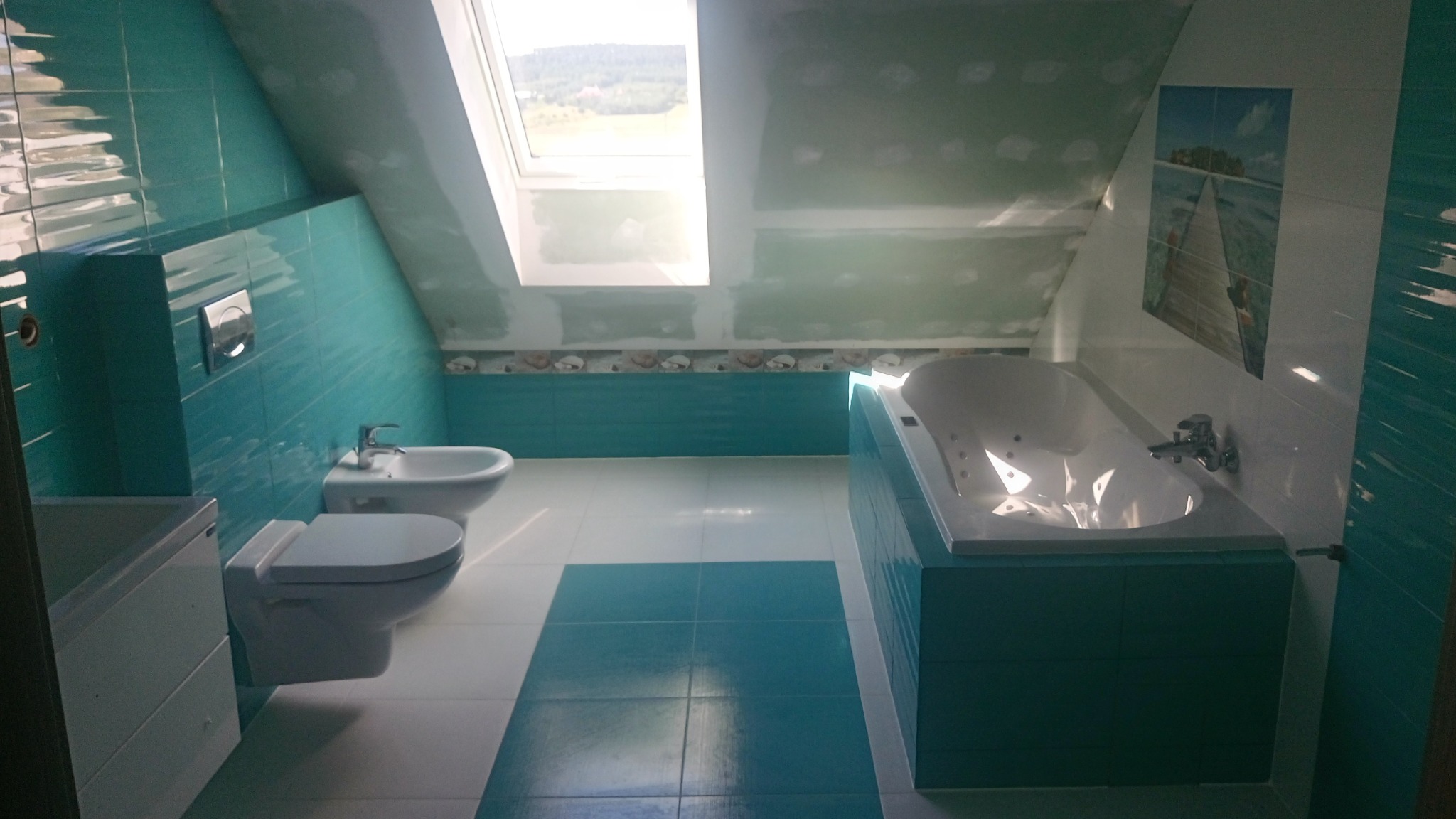 Turkusowo-biała łazienka na poddaszu ze skośnym oknem dachowym, widoczne płytki, wanna z hydromasażem, bidet i toaleta, ściany częściowo przygotowane do malowania.