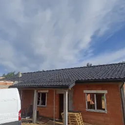 Dom w trakcie budowy z ciemnoszarą dachówką, widoczny fragment białego samochodu dostawczego, błękitne niebo z chmurami w tle.