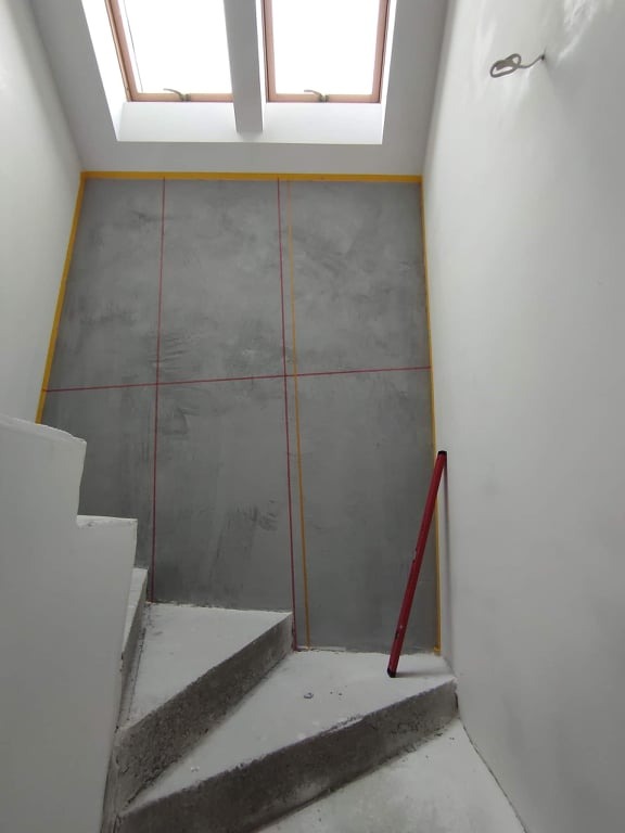 Ściana przy schodach przygotowana do wykończenia, z widocznymi liniami wyznaczonymi taśmą malarską i pionem, pod oknem dachowym.