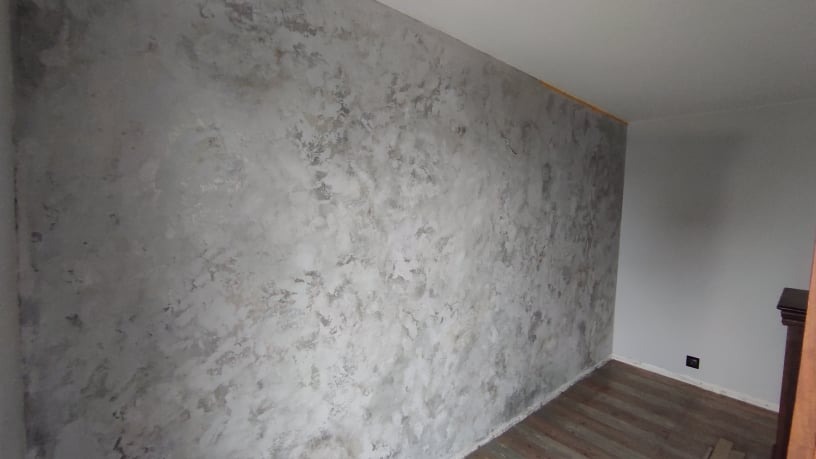Ściana w pokoju pokryta dekoracyjnym tynkiem w odcieniach szarości, imitującym efekt betonu architektonicznego, z widoczną fakturą i delikatnymi nierównościami.