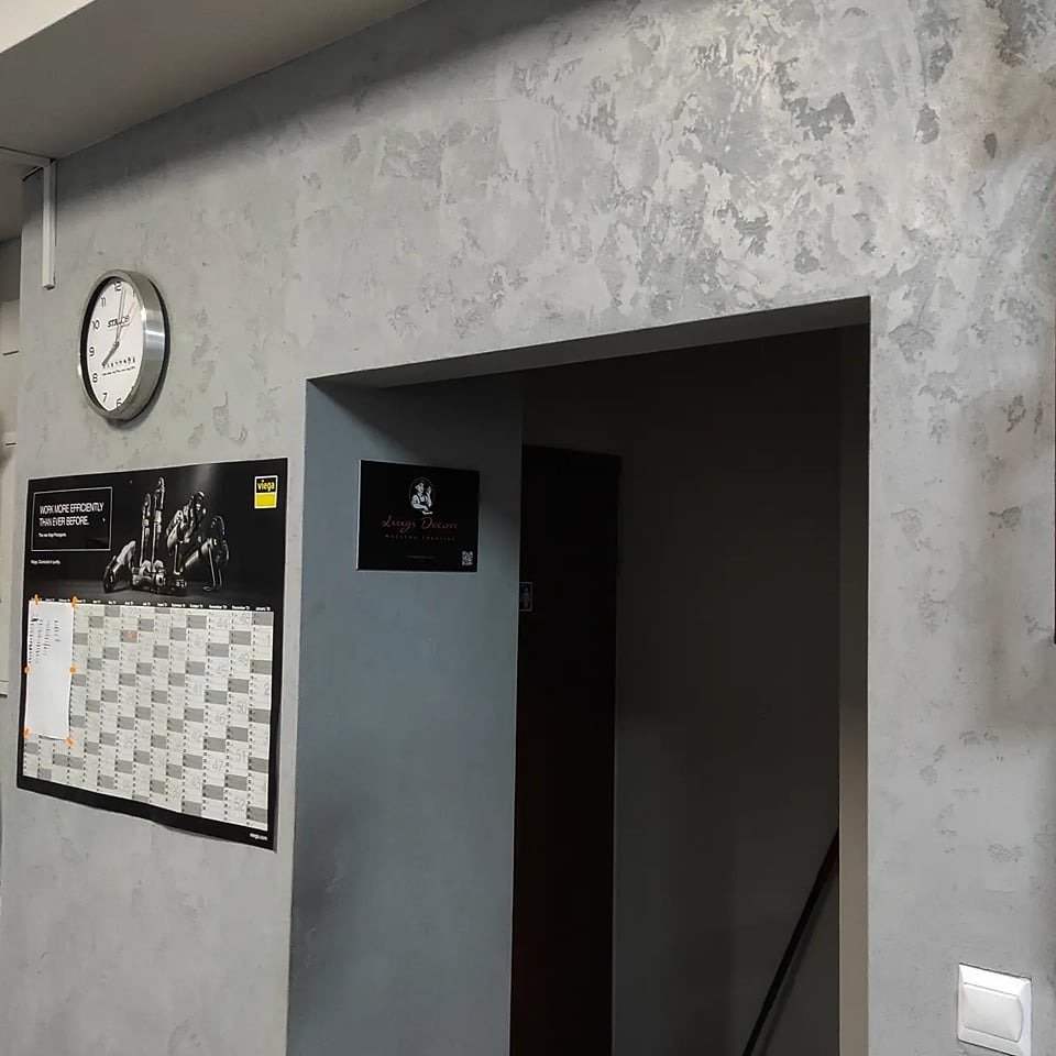 Ściana pokryta dekoracyjną szarą strukturą, widoczny zegar, kalendarz firmowy i mała tabliczka informacyjna obok wejścia do ciemnego pomieszczenia.