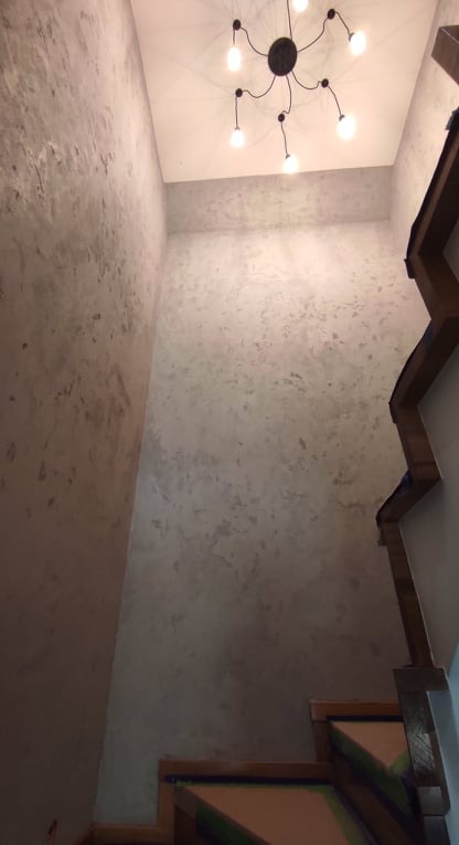 Ściana w korytarzu pokryta dekoracyjnym szarym tynkiem strukturalnym, drewniane schody z widocznymi zabezpieczeniami, nowoczesna lampa sufitowa z kilkoma żarówkami.