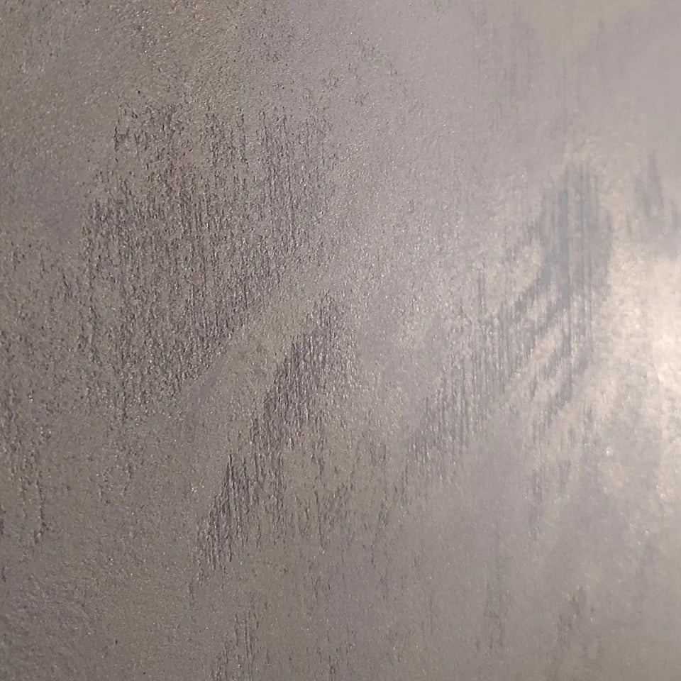 Ściana pokryta dekoracyjną, metaliczną farbą w odcieniach szarości z widoczną fakturą pionowych pociągnięć pędzla, tworząca efekt subtelnego cieniowania.