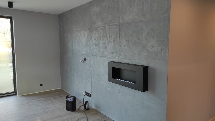 Nowoczesne wnętrze z szarą ścianą o fakturze betonu, na której wisi czarny biokominek, widoczne gniazdka elektryczne i przewody, jasna podłoga i okno z widokiem na zieleń.