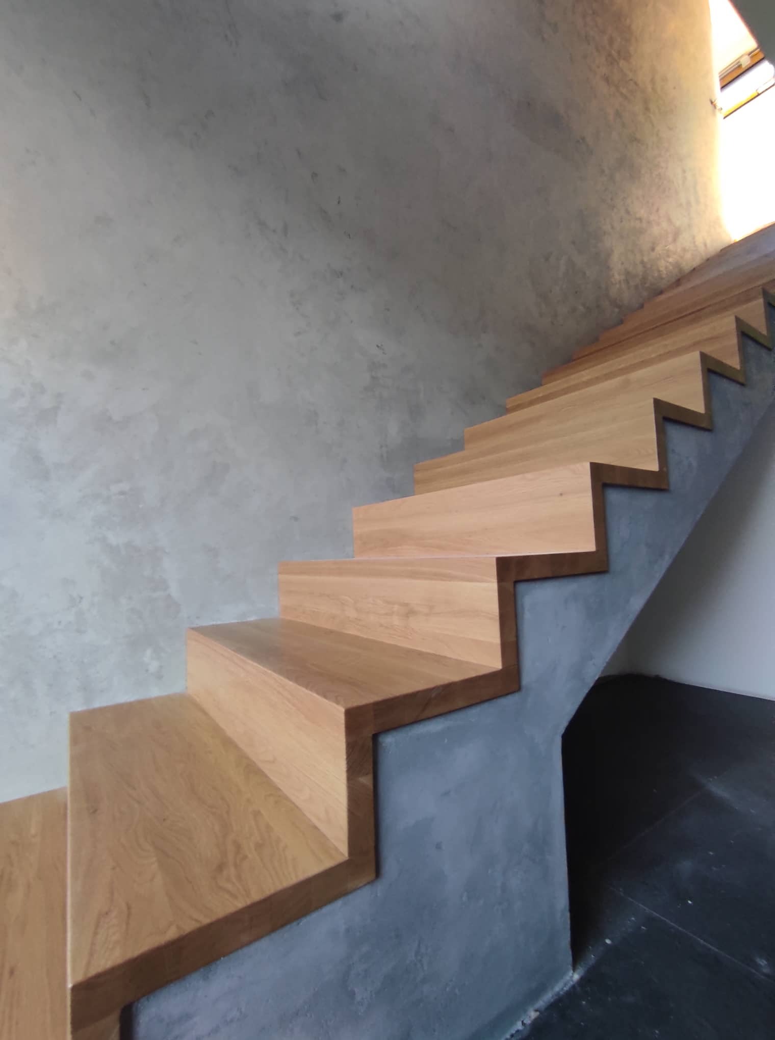Nowoczesne drewniane schody zintegrowane z szarą, betonową ścianą o surowym wykończeniu, oświetlone naturalnym światłem z góry.