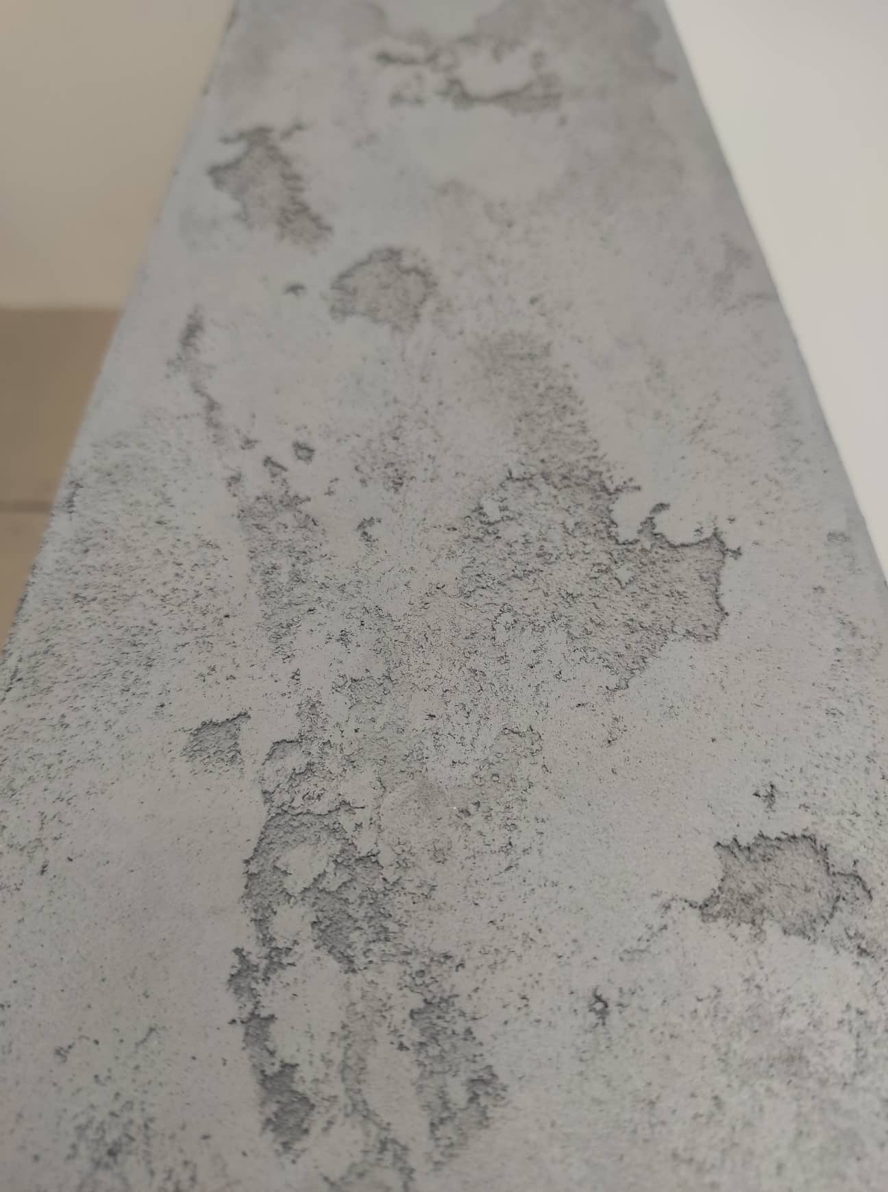 Fragment ściany z nierówną, szarą fakturą przypominającą beton architektoniczny, z widocznymi spękaniami i ubytkami.